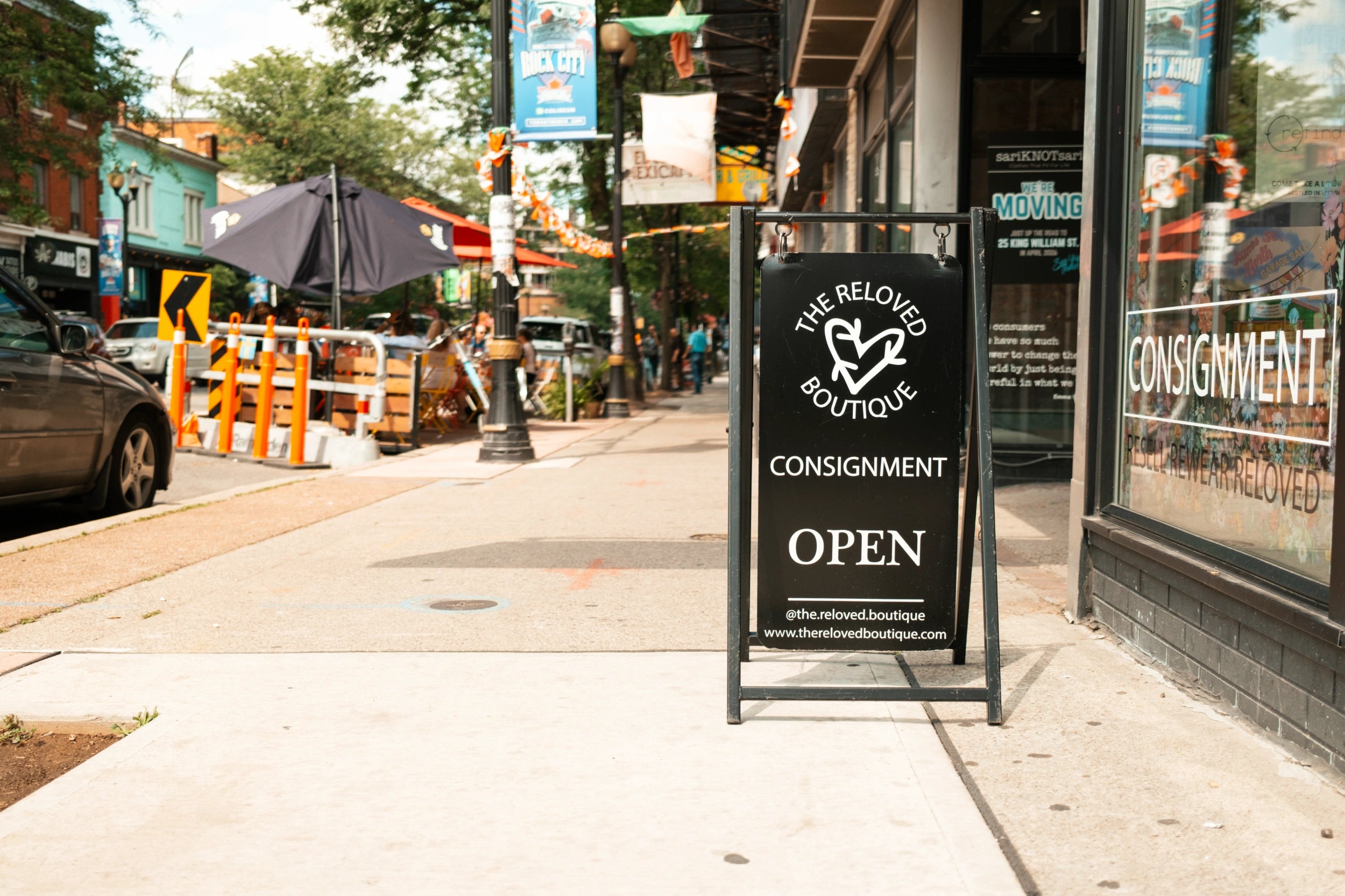A black A-frame sidewalk sign with The Reloved Boutique's logo on the sidewalk of James Street North in Hamilton.
