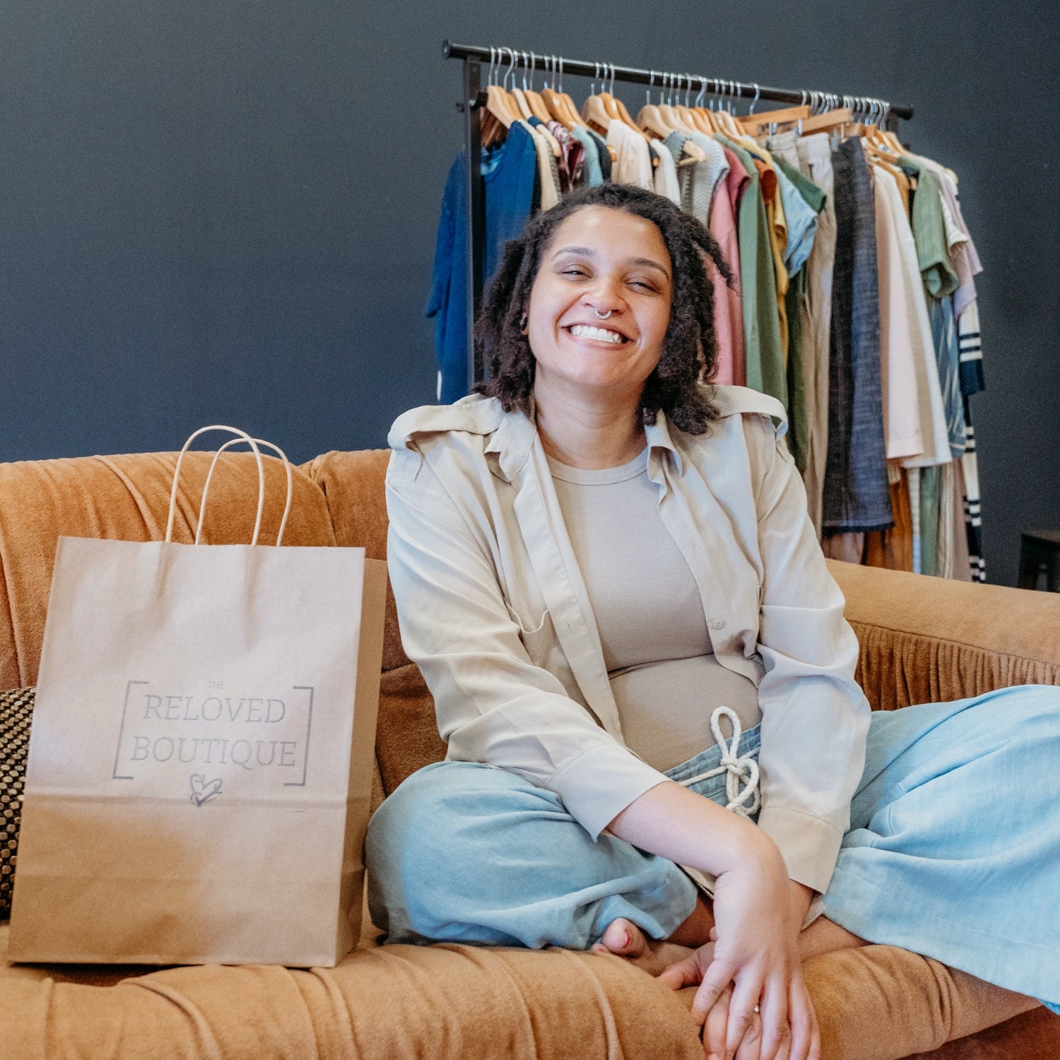 Lex of The Reloved Boutique sitting on a couch and smiling at the camera with a paper bag with the Reloved Boutique on it beside her.